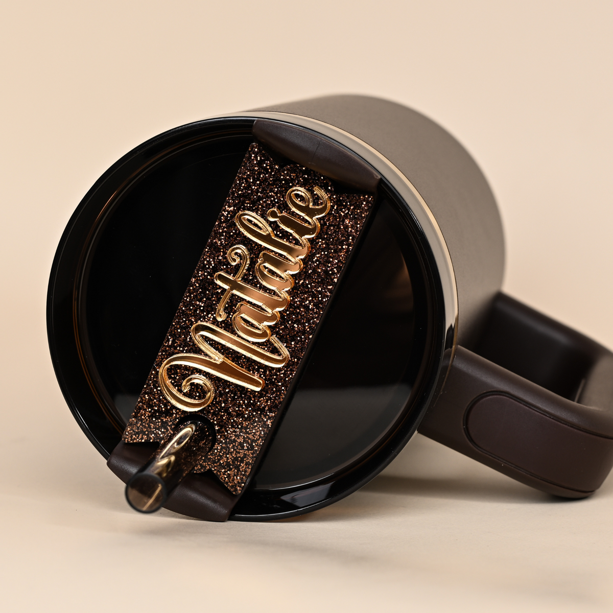 A chocolate gold stanley tumbler laying on it's side and a topper tag lays ontop with gold lettering and a glitter base. The Tag reads Natalie