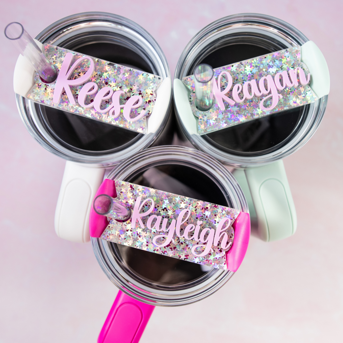 Three Stanley tumblers viewed from above, each topped with a sparkly glitter star acrylic name plate. The names Reese, Reagan, and Rayleigh are displayed in ballet pink, adding a charming personalized touch