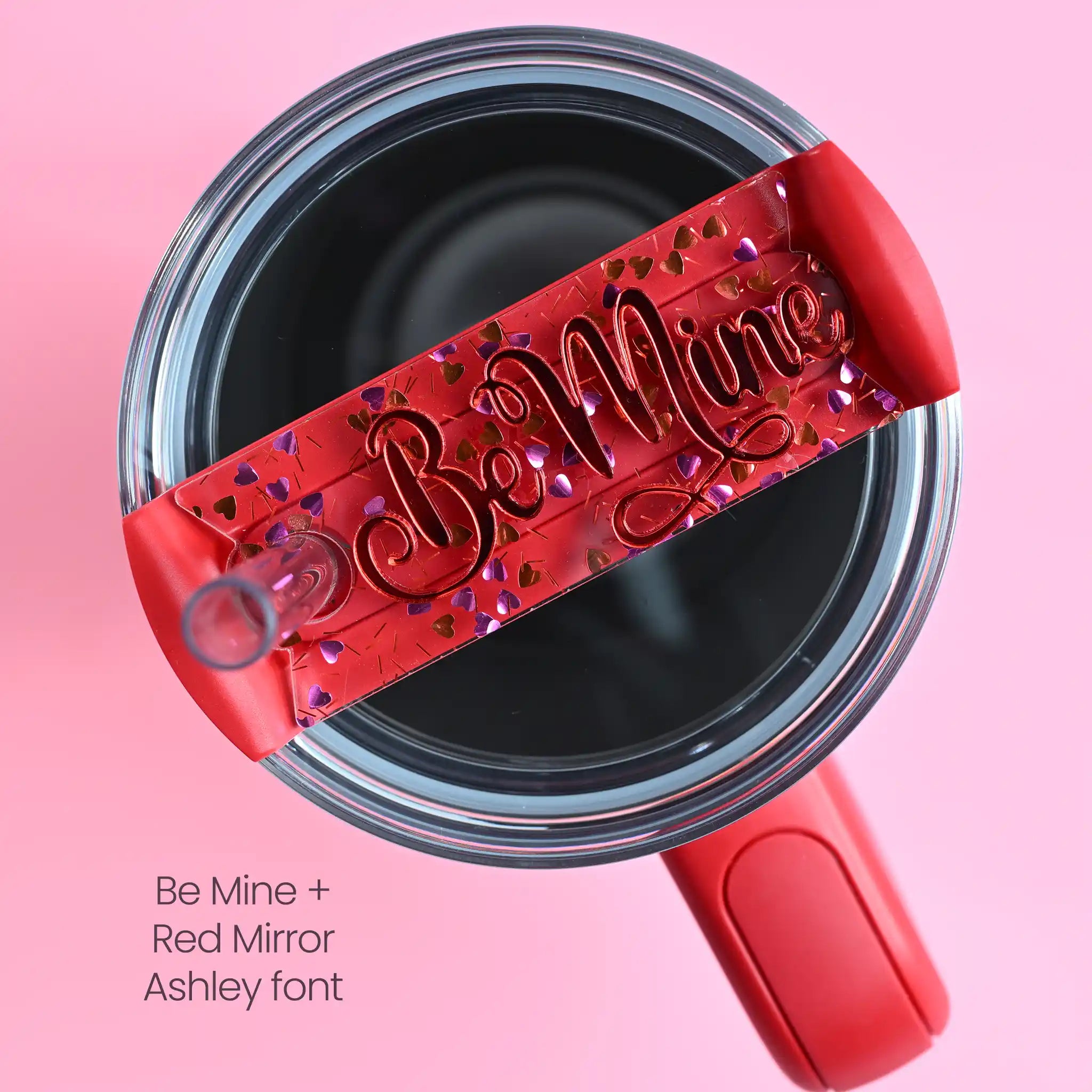 The top of a stanley tumbler shown with pink and red metallic foil hearts in clear acrylic with the text Be Mine written in a red mirror acrylic font in cursive letters.