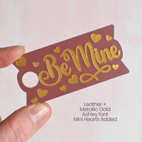 A single Topper Tag name plate in a burgundy/reddish brown semi-transparent acrylic with the text Be Mine written in metallic gold font with mini heart shapes surrounding the text.