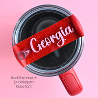 The top of a stanley tumbler shown with red shimmer acrylic with the name Georgia written in cursive in bubblegum pink acrylic font.